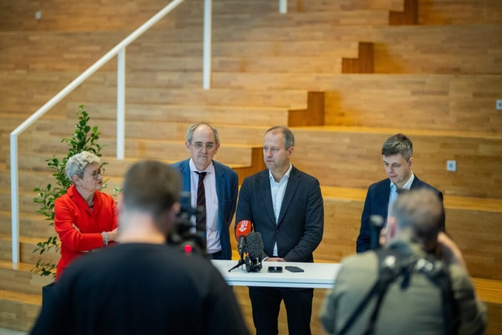 Four speakers stand at a high table with microphones at the press conference at ISM University while camera operators film them.