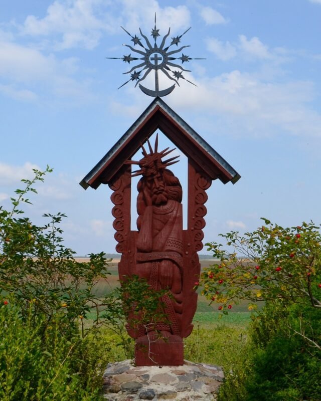 A carved wooden figure of Christ, seated with his head resting on his hand, under a small roofed shrine topped by a metal sun cross, in open fields.
